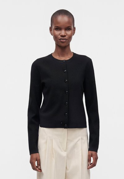 Woman wearing a black buttoned cardigan and light beige pleated trousers standing against a plain background.