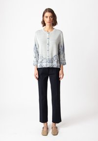 Light gray cardigan with blue floral pattern on hem and sleeves, round neckline, button closure, paired with black trousers and beige shoes.