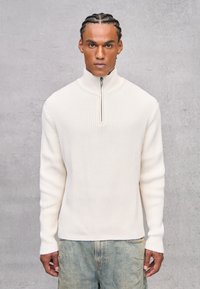 Maglione bianco a coste con colletto alto e metà zip, tessuto testurizzato, maniche lunghe e vestibilità comoda, abbinato a denim chiaro.