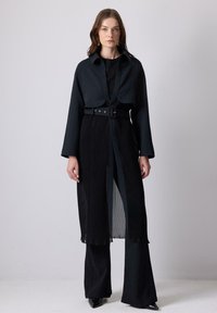 Black tailored coat with wide sleeves, pleated design, and a waist belt. Underneath, a fitted black top and flared pants complete the outfit.