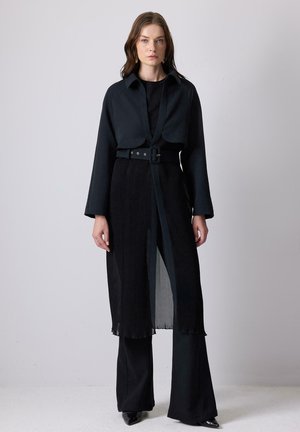 Abrigo negro ajustado con mangas anchas, diseño plisado y cinturón a la cintura. Debajo, una blusa negra ajustada y pantalones acampanados completan el conjunto.