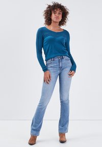 Turkusowy sweter z długim rękawem i dekoltem w serek, z teksturowanymi akcentami, w połączeniu z jasnoniebieskimi dzwonami i brązowymi botkami na obcasie.