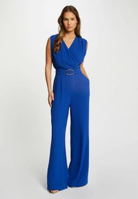 Jumpsuit bleu royal avec un décolleté croisé, sans manches, silhouette à jambes larges, ceinture avec quincaillerie en argent et poches latérales.