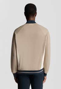 Beige sportsjakke med en marineblå krave og manchetter, der har en glat tekstur og et diskret farveblokdesign langs siderne.