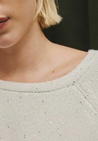 Close-up of a blonde person’s neck and shoulder wearing a light-colored, sequined knit sweater against a dark background.