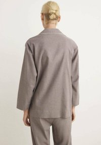Persona con cabello rubio recogido en un moño, vestida con una chaqueta gris de manga larga holgada y pantalones a juego, de pie de espaldas sobre un fondo liso.