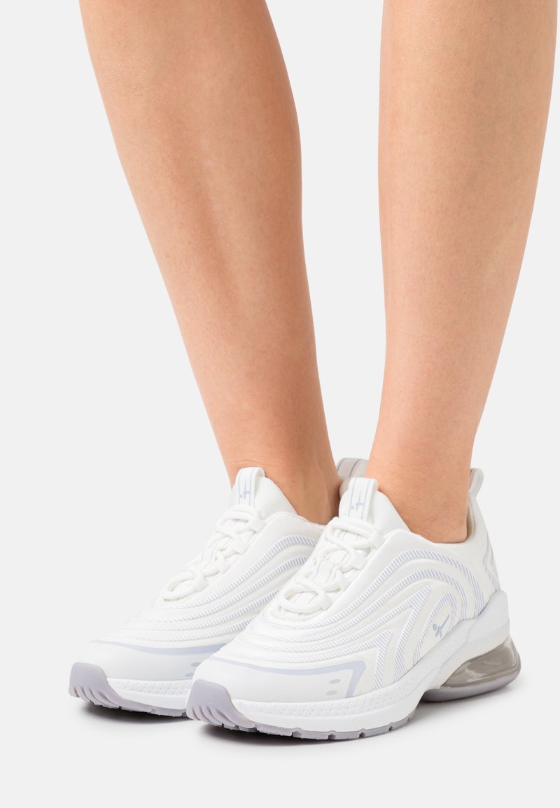 Personne portant des baskets blanches de sport avec un design texturé et un coussinet translucide au talon, debout sur un fond clair uni.