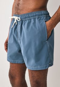 Plave kupaće hlače od lagane tkanine, s elastičnim pojasom koji ima vrpcu za vezanje, bočnim džepovima i kontrastnim šavovima.