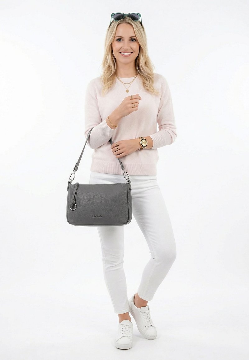 Mujer con suéter rosa claro, pantalones blancos y zapatillas blancas, sosteniendo un bolso gris con correa para el hombro, sonriendo y con gafas de sol en la cabeza.