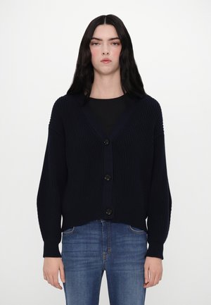 Marineblauwe ribbel cardigan met een V-hals, voorzien van vijf zwarte knopen en lange mouwen, gecombineerd met blauwe jeans voor een neutrale achtergrond.