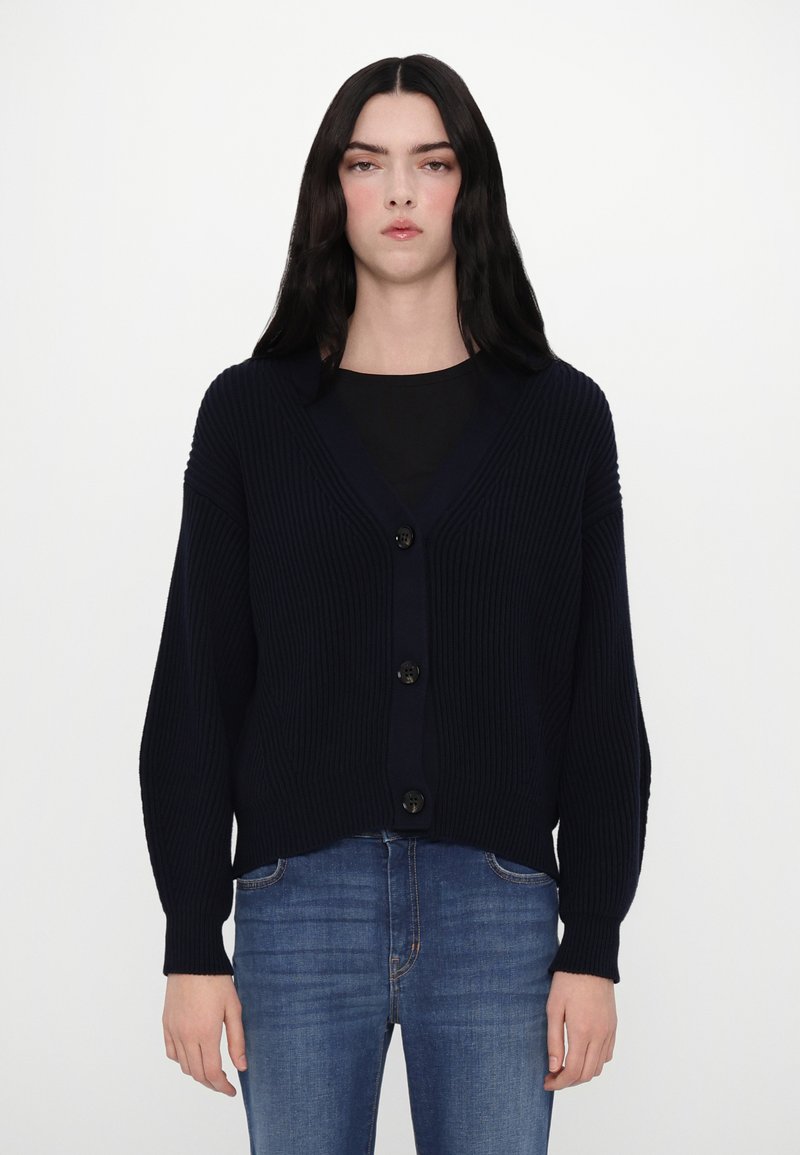 Marineblauer gerippter Cardigan mit V-Ausschnitt, ausgestattet mit fünf schwarzen Knöpfen und langen Ärmeln, kombiniert mit blauen Jeans vor einem neutralen Hintergrund.