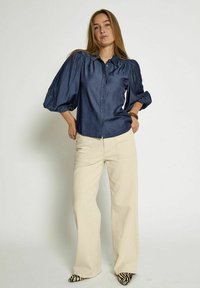 Chemise bleu marine à manches bouffantes ; présente une texture lisse. Associée à un pantalon ample beige clair et des chaussures à motif zèbre.