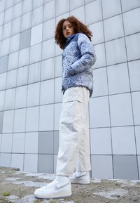 Jeune personne avec des cheveux bouclés portant une veste bleue à motifs, un pantalon cargo blanc et des baskets blanches Nike, se tenant à l'extérieur près d'un mur carrelé.