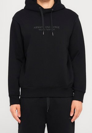 Czarna bluza z kapturem z napisem „Armani Exchange Milano/New York” na piersi, noszona przez osobę ze swobodnie opuszczonymi rękami przy bokach.