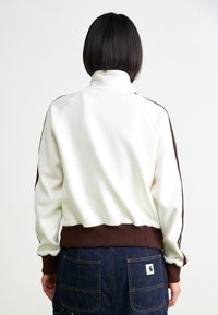 Vit sweatshirt med hög krage och bruna ribbade manschetter, med bruna sidostycken och en åtsittande midja, visad bakifrån.