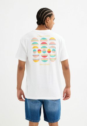 Camiseta blanca de manga corta con un patrón geométrico colorido en la parte posterior, con diseños circulares en rosa, amarillo y turquesa. Texto arriba y abajo.