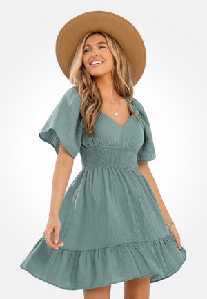 Uśmiechnięta kobieta w szerokim słomkowym kapeluszu i jasnoturkusowej sukience o fakturze z bufkami na rękawach, trzymająca rąbek sukienki, gdy się rozchyla.