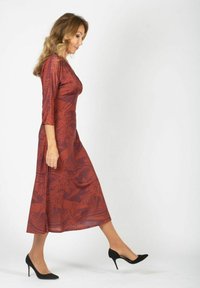 Kobieta z falującymi włosami spacerująca w midi sukience w kolorze rdzy w wzory oraz czarnych szpilkach na tle jednolitej białej ściany.