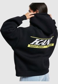 Sort hætte sweatshirt med hvid og neongul tekst "KAN NIGHT RIDER EST 1989" på ryggen, med en afslappet pasform og ribbestrikkede manchetter.