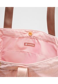 Růžová látková taška s hnědými popruhy. Podšívka obsahuje malou kapsu a štítek s logem. Viditelný detail s drukem.