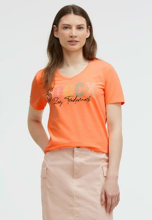 Junge Frau mit langen braunen Haaren, die ein orangefarbenes Kurzarmshirt mit mehrfarbigem "SOCCX"-Text und einen hellrosa Rock trägt.