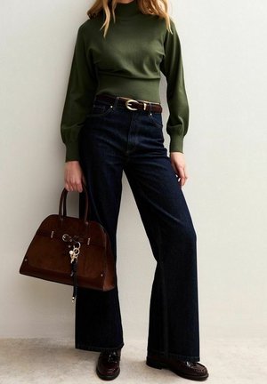 Femme portant un haut vert à manches longues et un jean large foncé tient un grand sac à main marron, debout contre un mur clair uni.