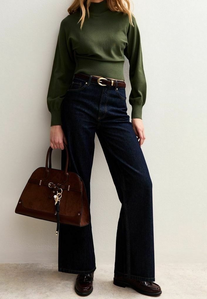 Femme portant un haut vert à manches longues et un jean large foncé tient un grand sac à main marron, debout contre un mur clair uni.