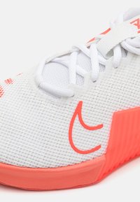 Zapatilla deportiva blanca de malla con detalles en naranja, que presenta una suela texturizada, parte superior perforada y un diseño de logotipo contrastante en el lateral.