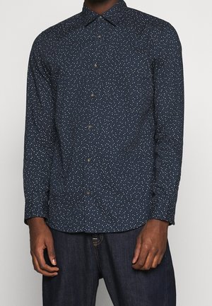 Homme portant une chemise bleu marine à manches longues avec un petit motif à pois blancs, associée à un jean bleu foncé.