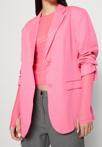 Růžový blazer vyrobený z hladké látky, s jedním knoflíkem, klopovými límci a ohrnutými rukávy. Nosený přes odpovídající top.
