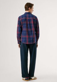 Camisa de cuadros en azul marino con líneas rojas y verdes, puños con botones y dobladillo curvado. Combinada con pantalones oscuros de corte holgado y zapatillas multicolor.