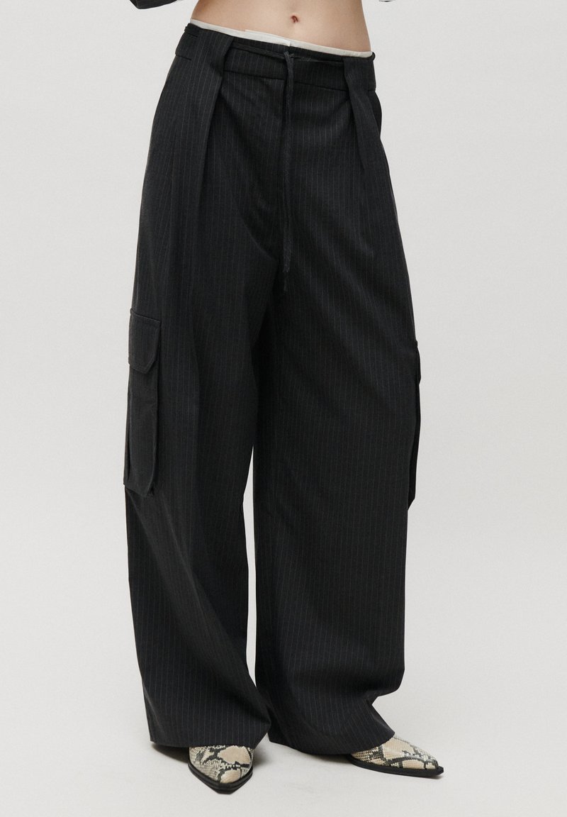 Pantalon noir à large jambe à fines rayures avec poches cargo latérales, porté avec des chaussures pointues en cuir de serpent sur fond blanc.