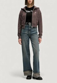 Felpa con cappuccio marrone con zip e tasche, abbinata a una maglia nera a coste con logo bianco. Jeans blu vita alta con gambe larghe e risvolti a contrasto.