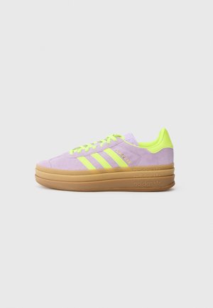 Sneaker Adidas Gazelle na niskiej podeszwie z pastelowofioletowego zamszu z jaskrawożółtymi paskami, sznurowadłami i wnętrzem, na prostym białym tle.