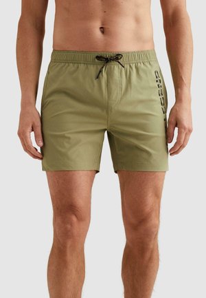 Mužský trup a nohy oblečené v olivovo zelených plavkových šortkách s čiernym šnúrkou a vertikálnym čiernym textom na pravej stehne, stojaci na neutrálnom pozadí.