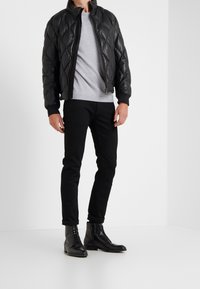 Blouson aviateur matelassé en cuir noir, chemise grise à col rond, jean slim noir et bottes noires à lacets, debout devant un fond blanc.