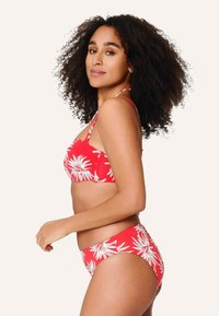 Červené bikiny s bielym kvetinovým vzorom, s tenkými ramienkami a štruktúrovaným vrchom. Vysoký pás na spodnej časti. Model pózuje v bočnom profile.