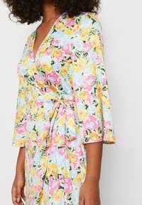 Blommig omlottklänning i pastellfärger. Har klockärmar, midjeband och ett livligt blommönster på en ljusblå bakgrund.