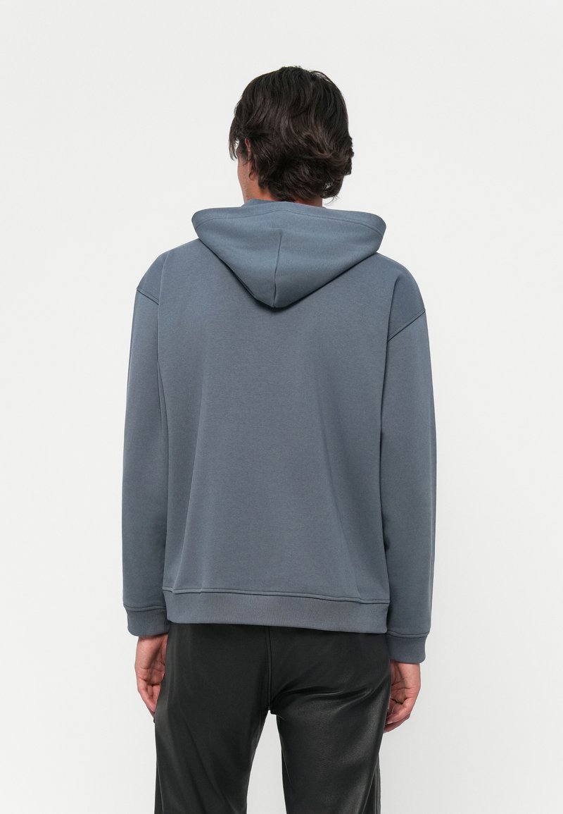 Hombre con cabello oscuro que lleva una sudadera azul y pantalones negros, visto desde atrás sobre un fondo claro y liso.