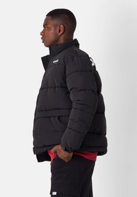 Giacca a vento nera con colletto alto, motivo trapuntato, zip frontale e dettaglio del logo. Indossata sopra una maglietta rossa, abbinata a pantaloni cargo.