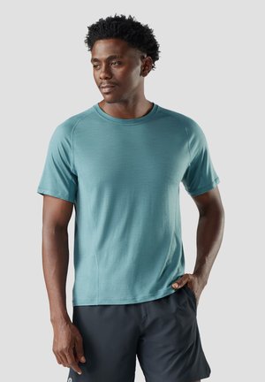 Uomo con capelli ricci che indossa una camicia a maniche corte azzurro chiaro e pantaloncini scuri, in piedi con una mano in tasca davanti a uno sfondo neutro.