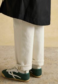 Biele rollované džínsy v kombinácii so zelenými teniskami s bielymi akcentmi a gumovou podrážkou. Čierna bunda čiastočne zakrýva džínsy.