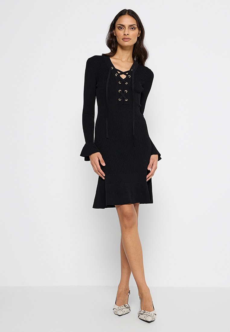 Kobieta stoi na białym tle, ubrana w czarną, długą dzianinową sukienkę z dekoltem w sznurowanie i rozszerzanymi mankietami, w zestawieniu z białymi butami na obcasie w wzory.