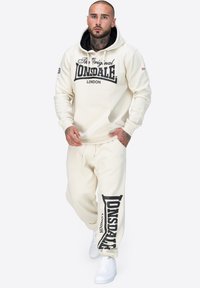 Sudadera con capucha y pantalones de chándal en color crema, con el texto negro "LONSDALE" y logo. Material de algodón suave, corte relajado, bolsillo frontal y capucha con cordón.