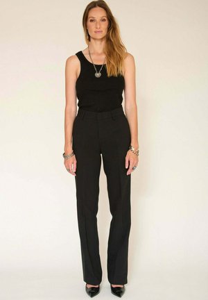 Débardeur noir associé à un pantalon noir ajusté, en tissu lisse, avec des plis subtils. Des chaussures noires à bout pointu complètent le look.