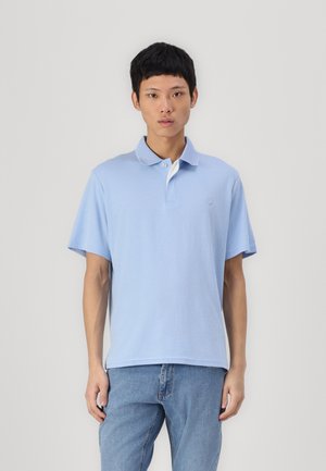 Light blue polo shirt with a soft texture, collar, and white accents. The model wears it with blue jeans, displaying a relaxed fit.