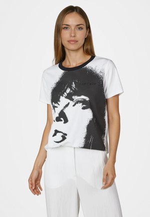 Frau mit langen Haaren, die ein weißes T-Shirt mit einer großen schwarz-weißen Grafik eines Frauenkopfs und weiße Hosen trägt, vor einem schlichten Hintergrund stehend.