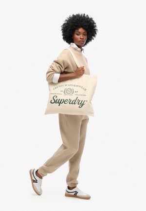 Femme portant un survêtement beige et des baskets blanches, tenant un sac fourre-tout beige avec le logo "Superdry Premium Sportswear" en lettres vertes.