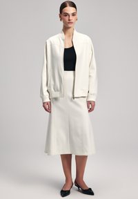Giacca bomber bianca con polsini a costine, abbinata a un top nero aderente e a una gonna midi a campana color crema. Tacchi a punta nera completano l'outfit.