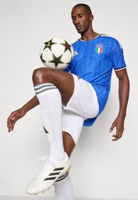 Plava nogometna majica s teksturiranim dizajnom, bijele kratke hlače i čarape sa prugama. Drži uzorak lopte za nogomet. Bijeli kopački s crnim detaljima.